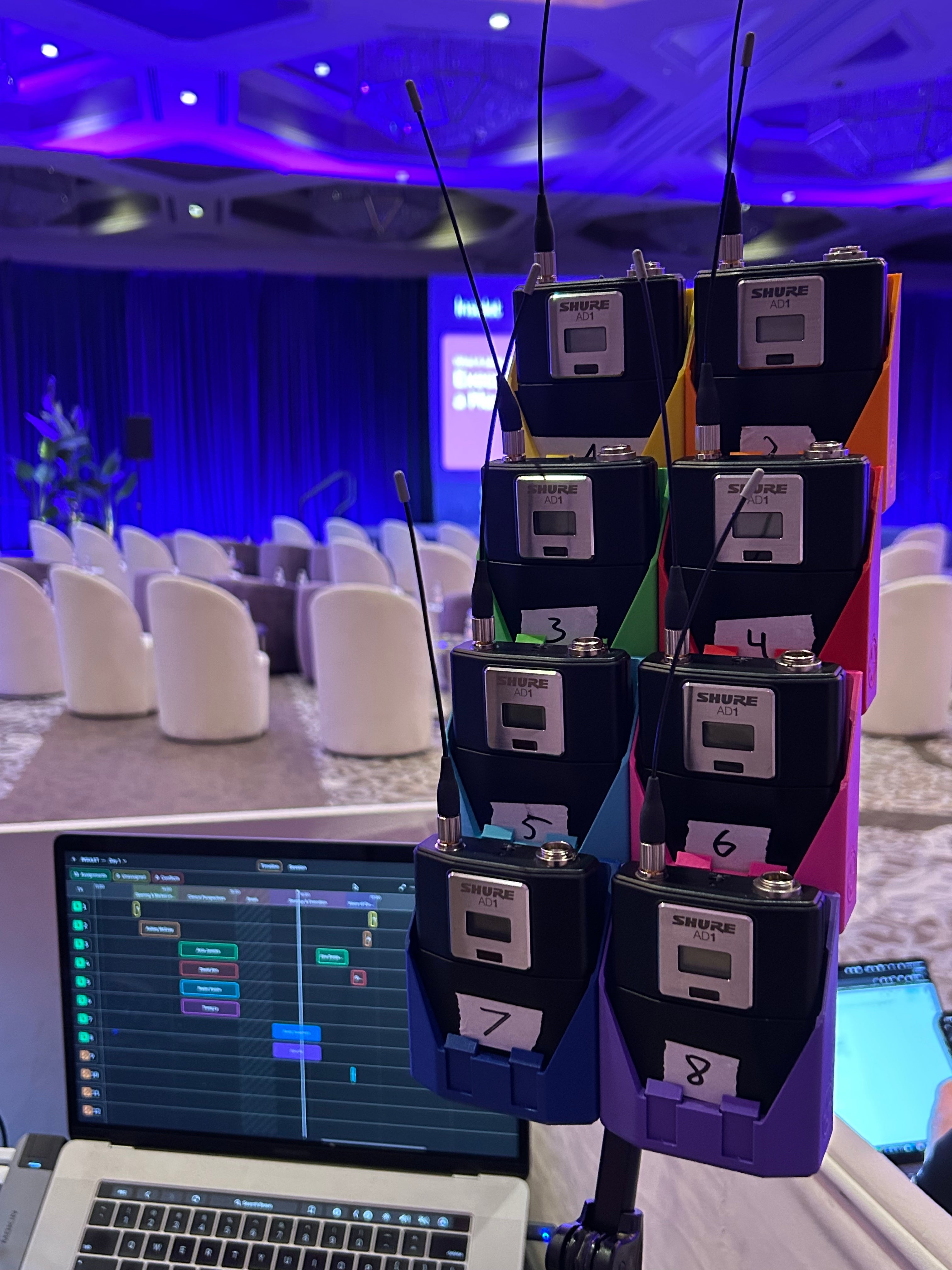 Eight color-coded StageStrike packHolders stacked on a packBase, numbered 1 to 8 and holding Shure AD1 wireless bodypacks, beside a laptop running micAddy at a corporate event.