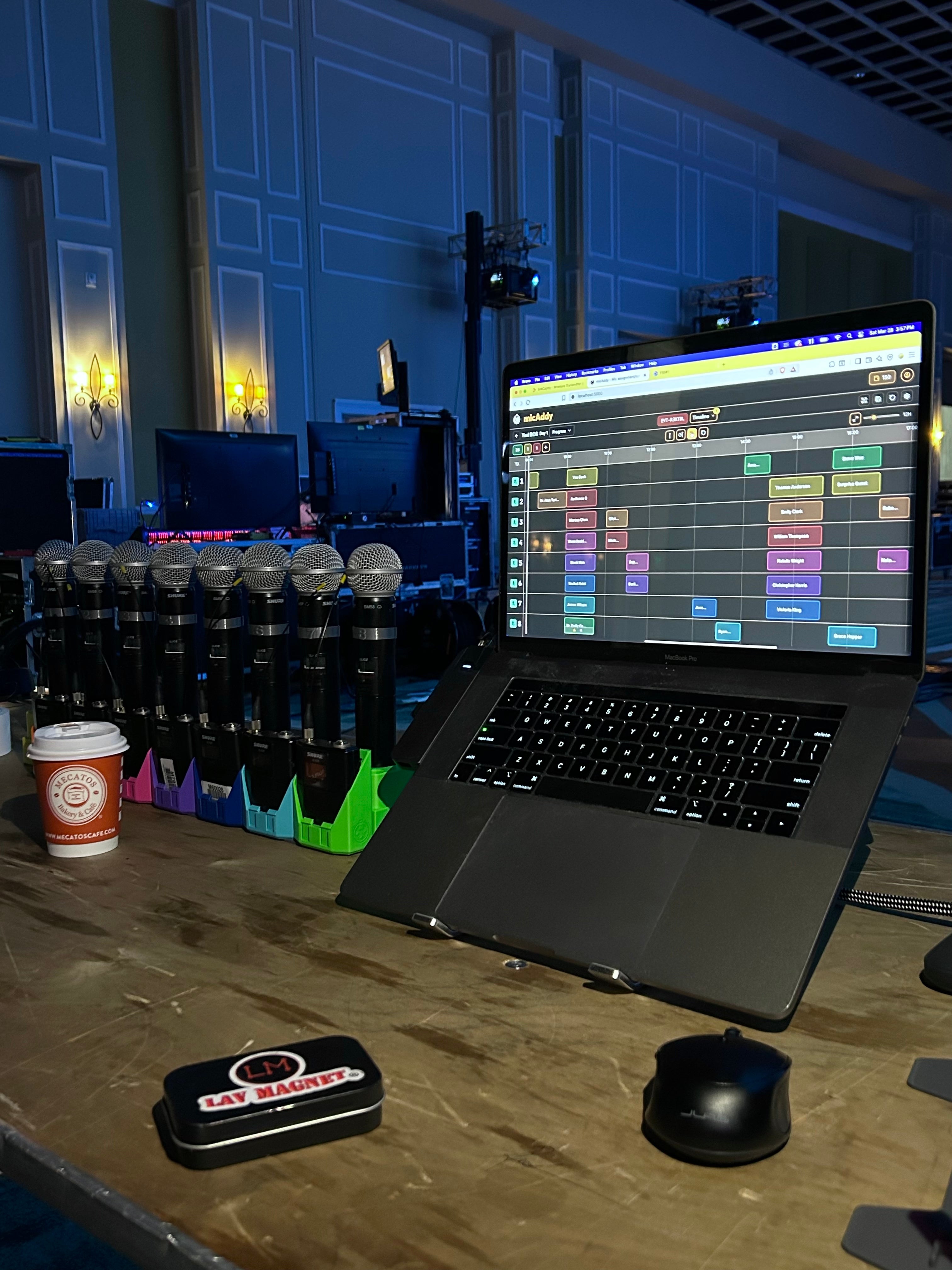 A row of color-coded StageStrike miQbs holding wireless handheld microphones next to a laptop running micAddy, the on-screen channel colors matching the holders.