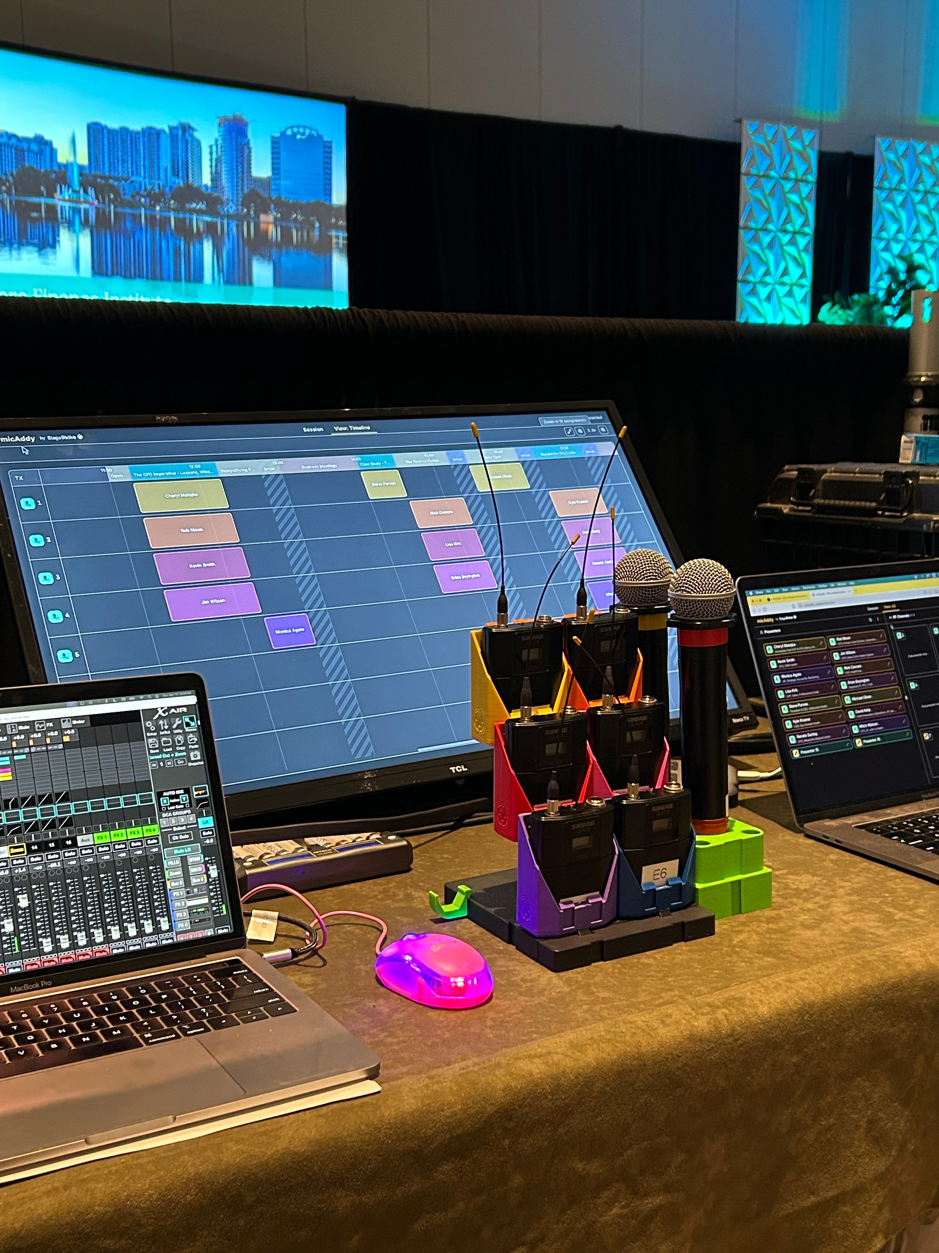 Corporate audio tech table with color-coded StageStrike packHolders and wireless handheld mics on a packBase, a mixing console, and micAddy's color-matched assignment timeline on the monitor.